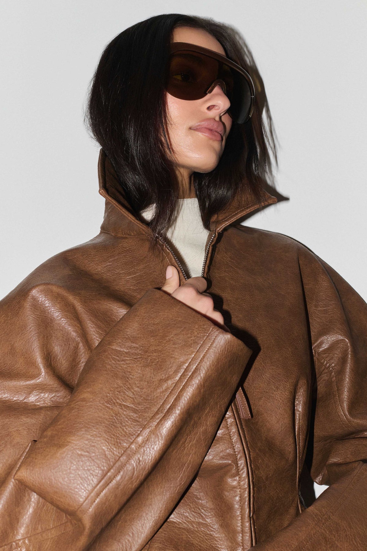 A model wearing an oversized brown faux-leather jacket with a high collar and zipper, styled with dark sunglasses and a cream turtleneck against a light gray background.