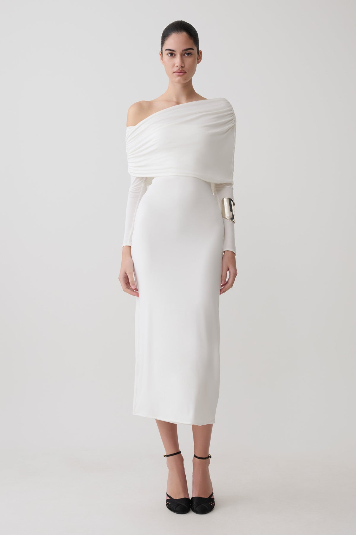 A woman modeling a long off-the-shoulder white dress with a draped wrap detail, holding a textured gold clutch, against a plain light gray studio background.