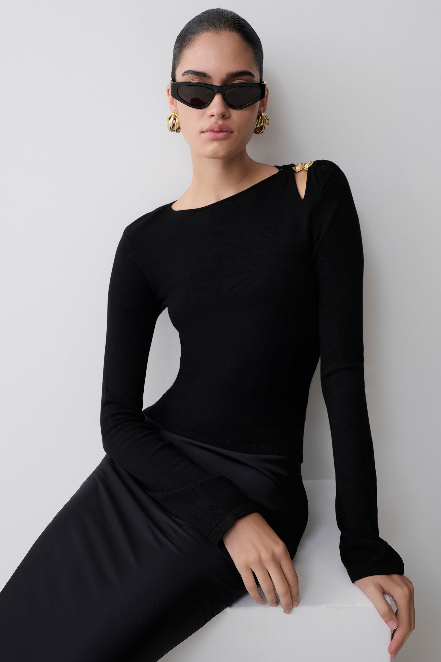 Woman wearing a black long-sleeve dress and sunglasses against a white background.