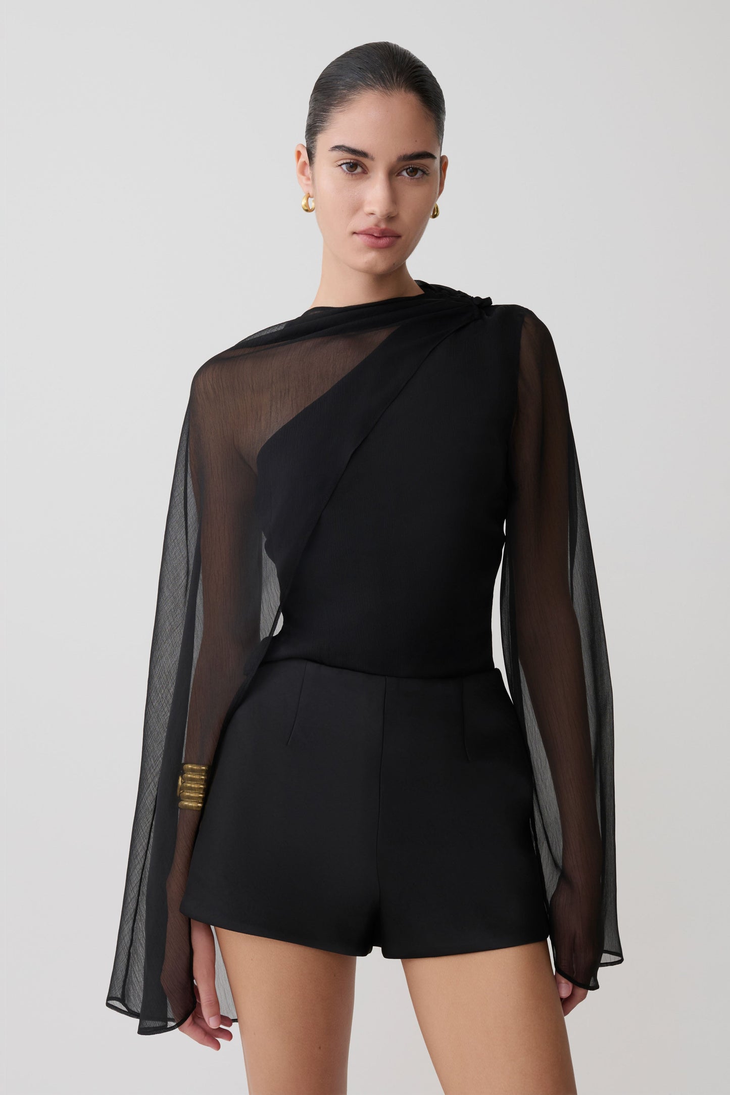 Woman wearing a black outfit with sheer sleeves on a plain background.