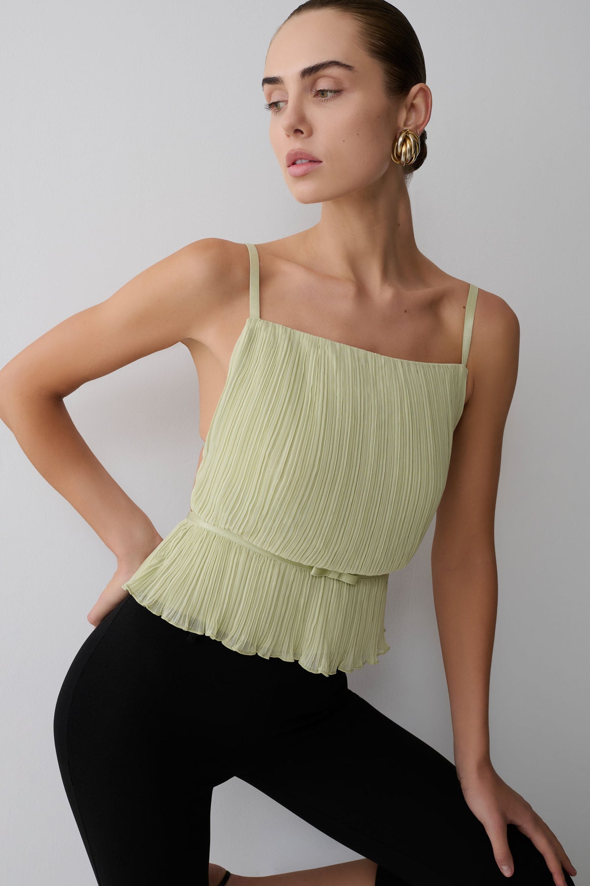 Woman wearing a light green pleated top and black pants against a plain background