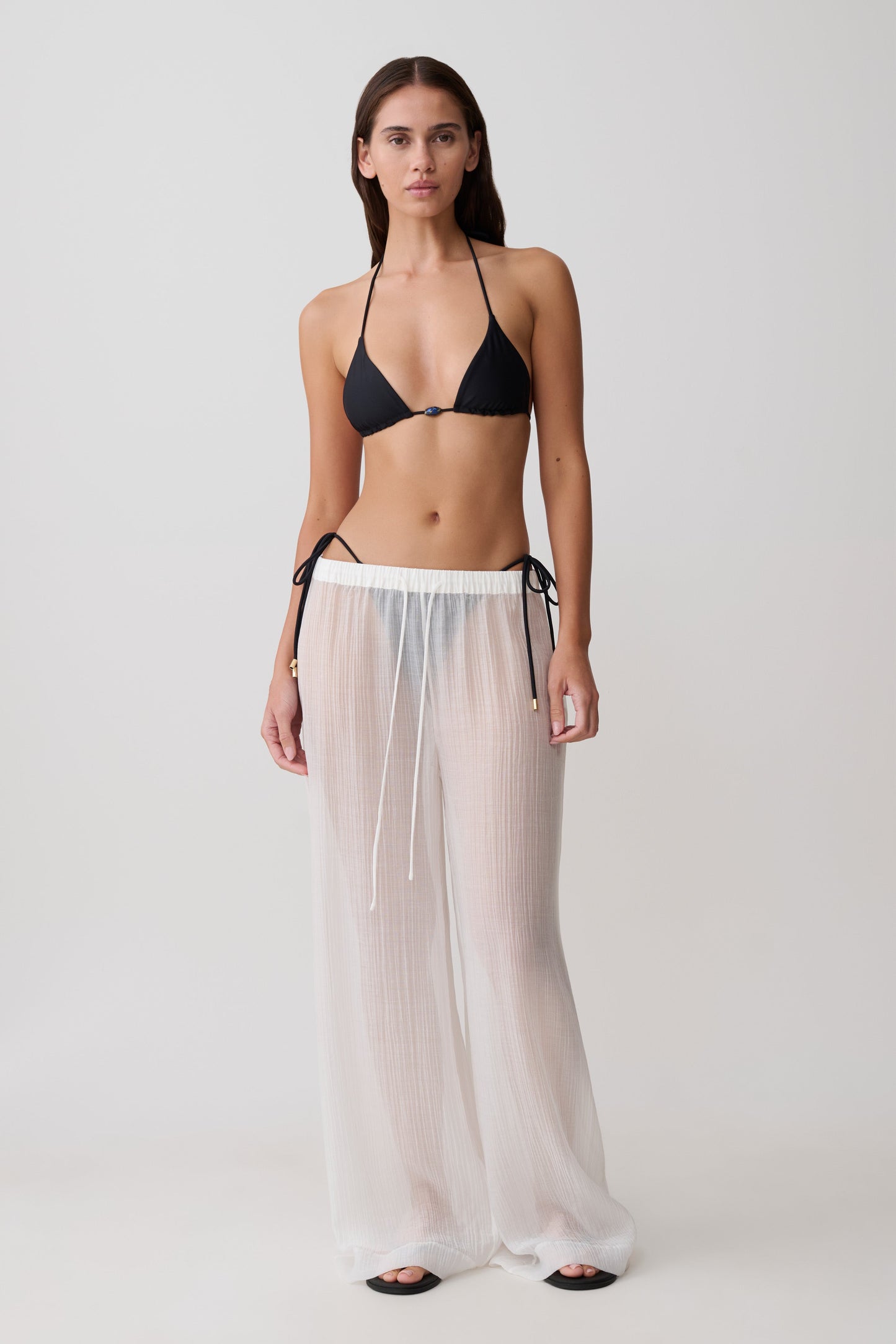 Woman wearing a black bikini top and sheer white drawstring wide pants.