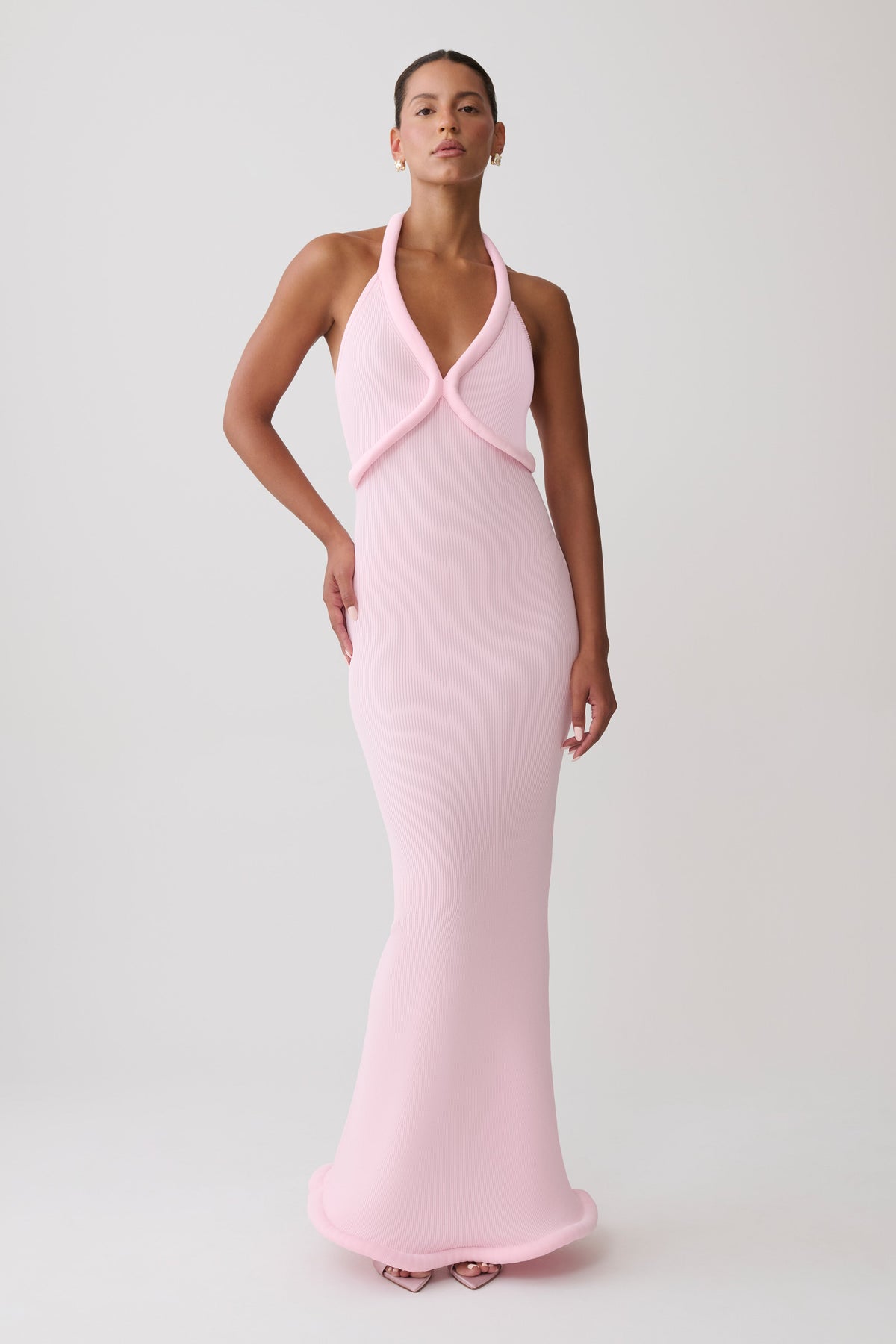 A woman modeling a light pink halter-neck maxi dress with a wrap-style bodice, photographed against a plain light gray studio background.