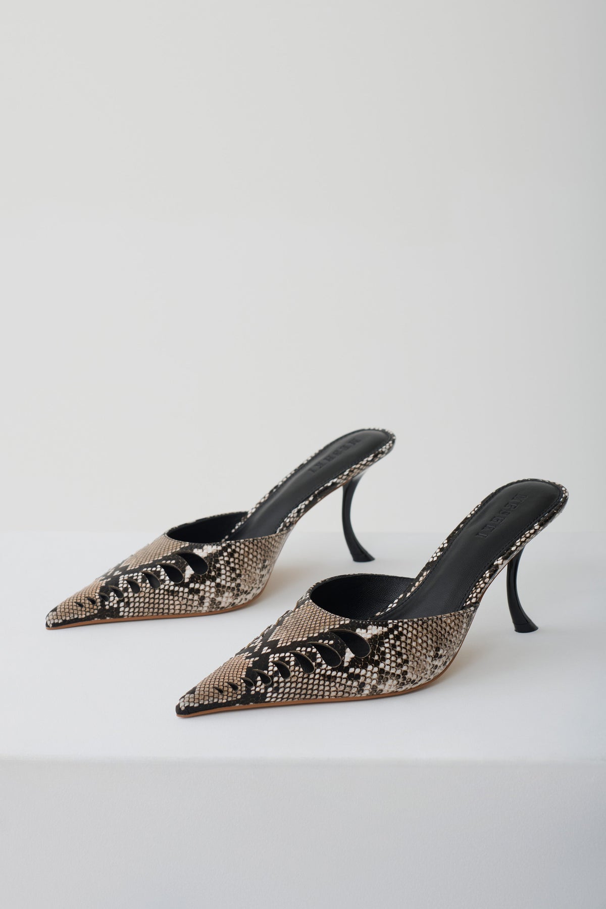 A pair of pointed-toe mule heels with a black and beige snakeskin pattern, displayed on a clean white studio background.