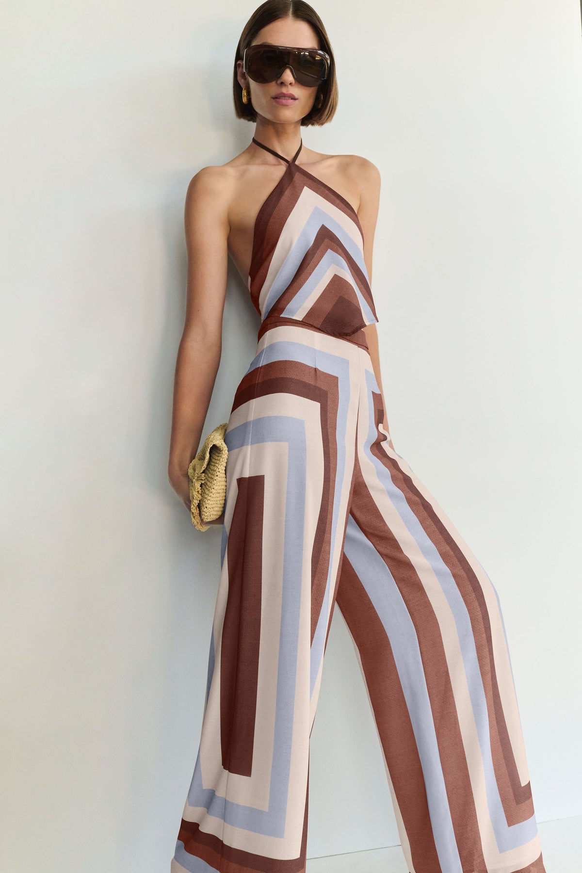 A woman modeling a sleeveless halter-neck top and wide-leg pants with a geometric striped pattern in brown, beige, and light blue, accessorised with dark sunglasses and a woven clutch against a plain light background.