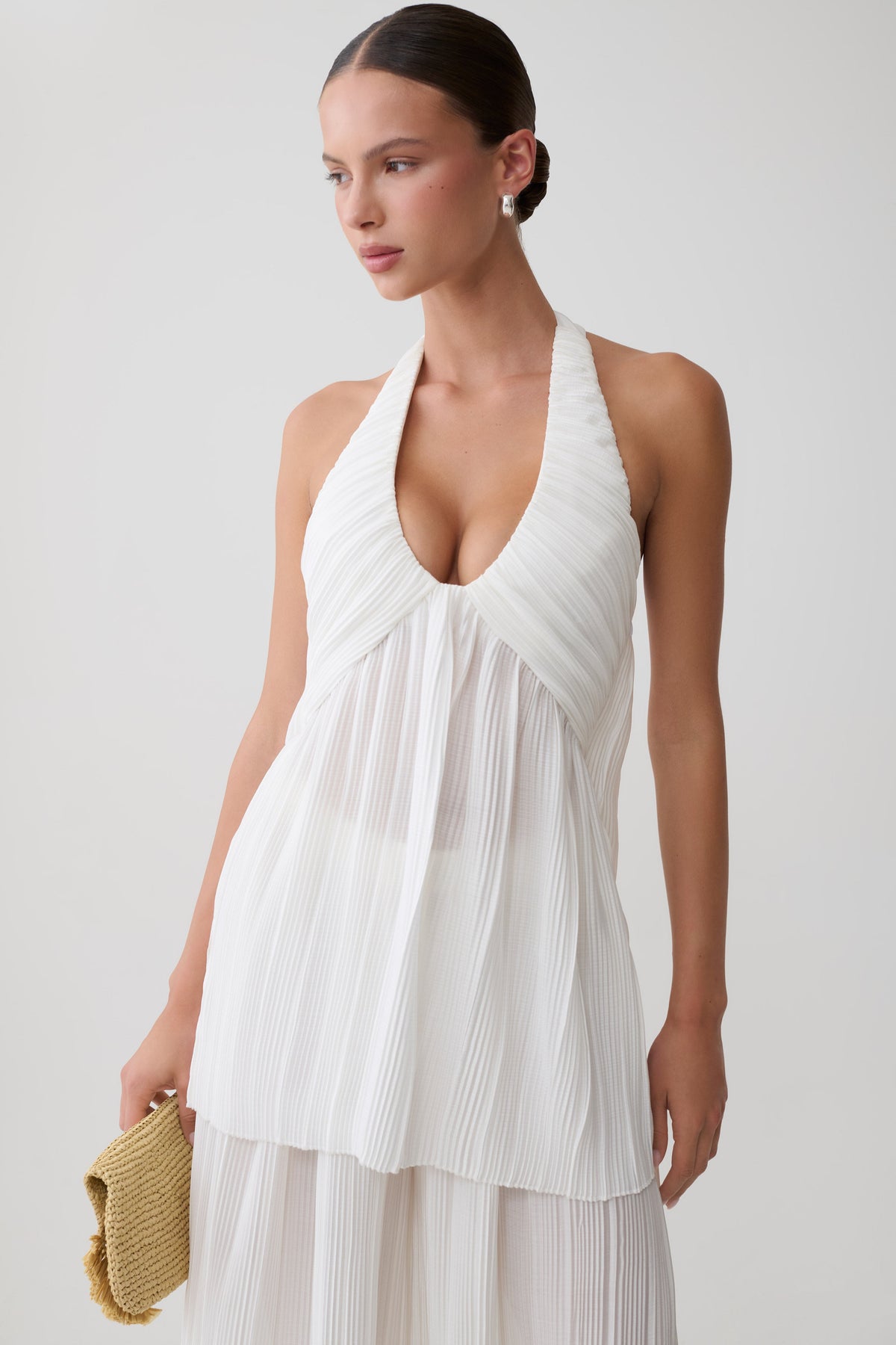 A woman modeling a white sleeveless halter-neck dress with a deep V-neckline and pleated texture, holding a small woven clutch against a plain light gray studio background.