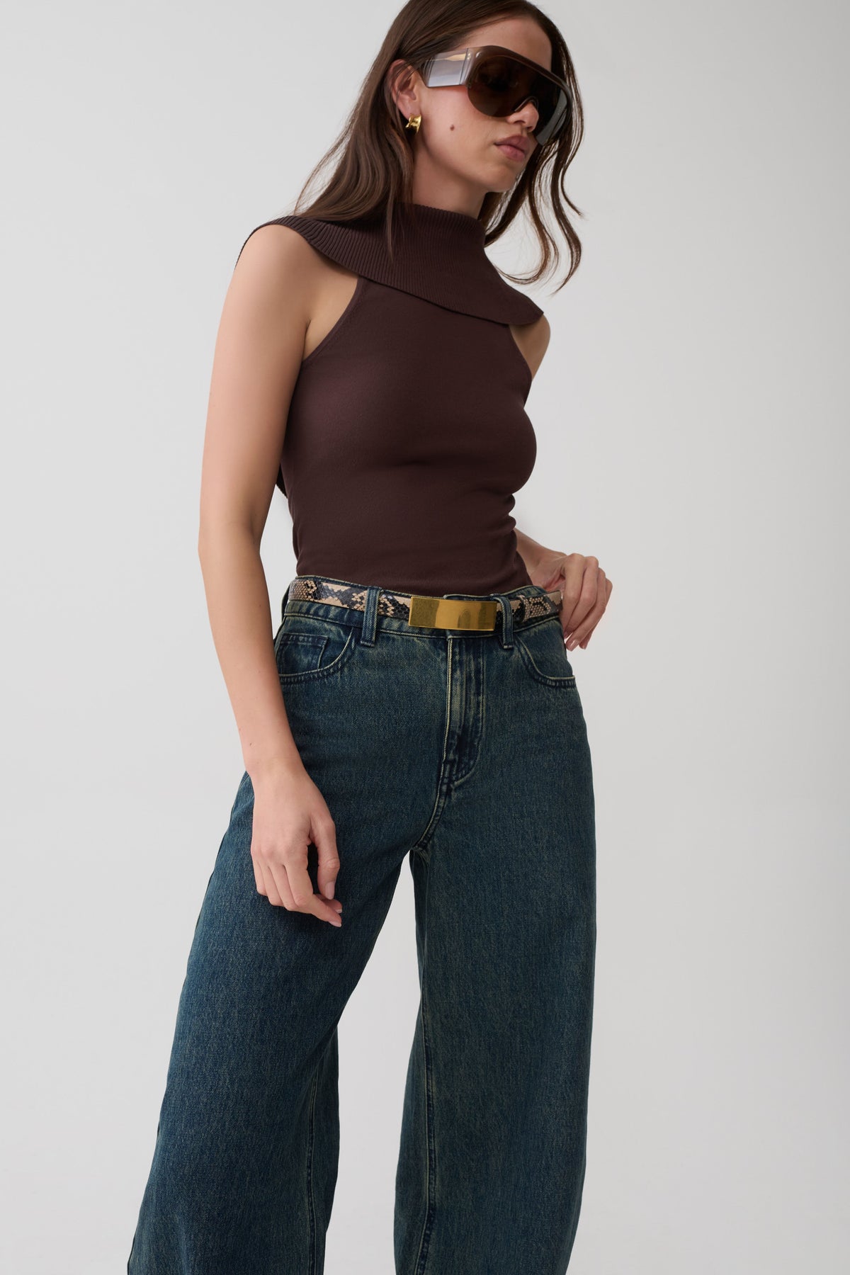 A woman modeling a sleeveless dark brown mock-neck top paired with dark blue wide-leg jeans and a gold belt, set against a plain light gray studio background.