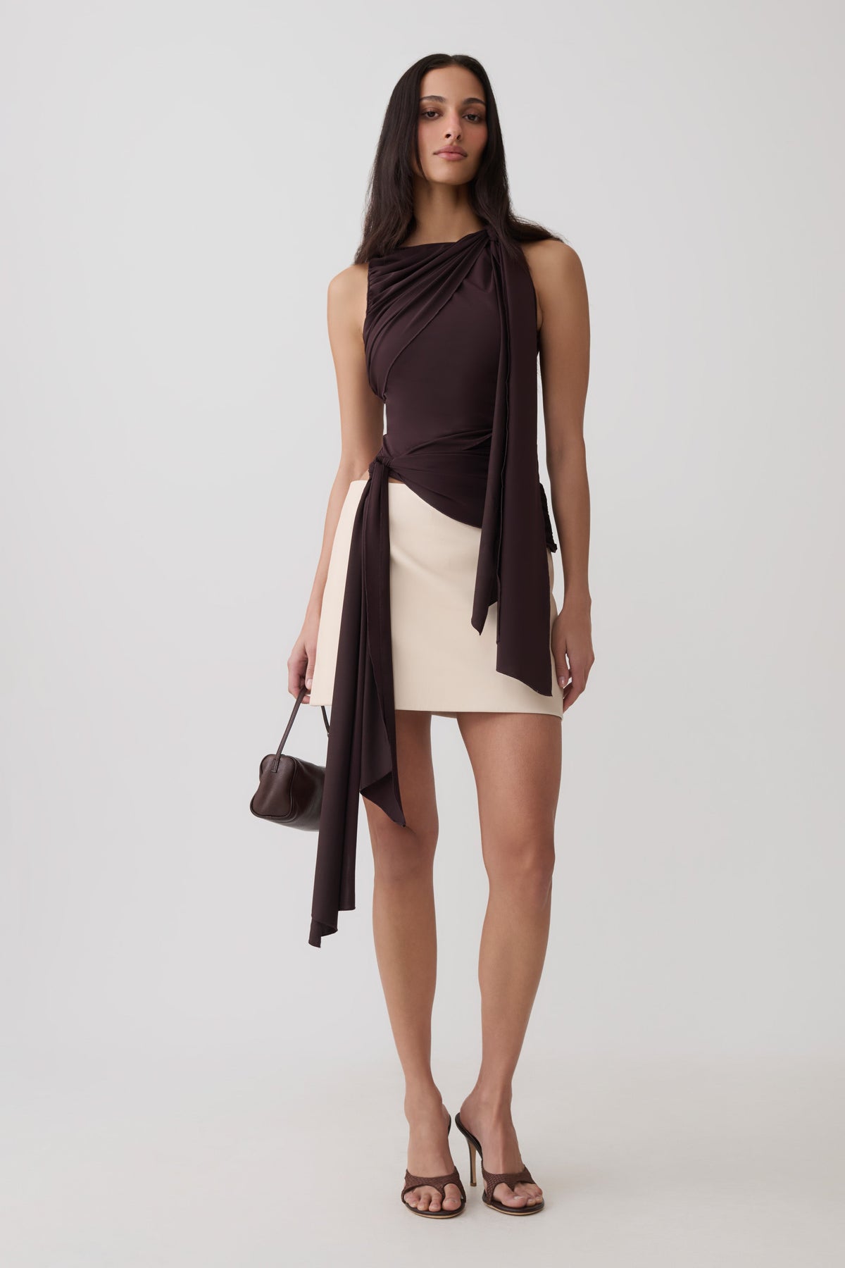 A woman models a sleeveless draped top in dark brown with a cream-colored mini skirt, holding a small dark brown handbag against a light gray studio background.