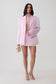 A model stands against a light gray studio background wearing a light pink tailored blazer over a matching pink mock-neck top and mini skirt. She is holding a small structured cream handbag and wearing clear high-heel sandals.