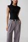 A model wearing a black sleeveless textured top and high-waisted gray pleated trousers with a black belt featuring a silver buckle, shot against a light gray studio background.