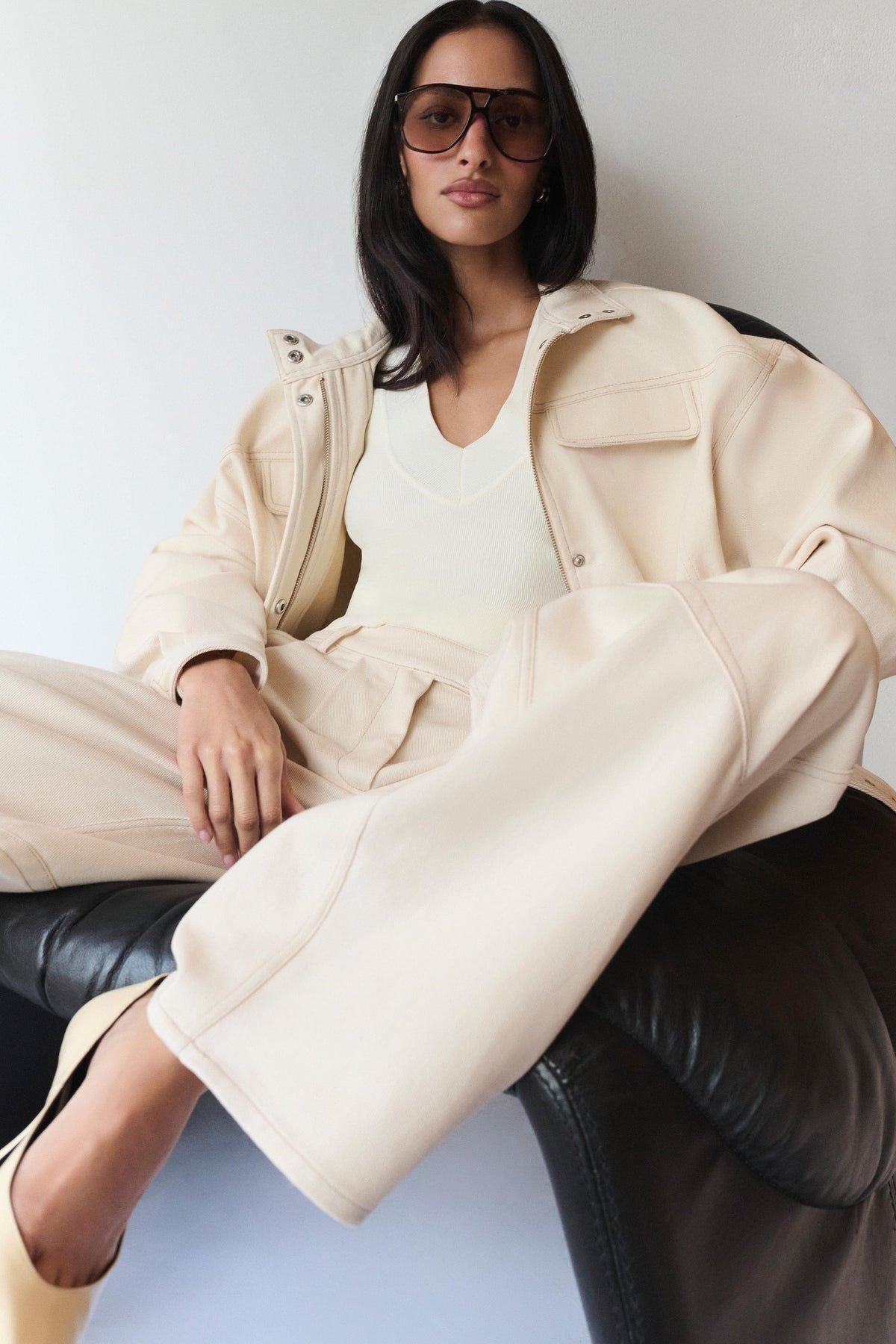 A woman wearing a cream-colored oversized denim jacket, matching wide-leg denim pants, and a white top, sitting on a black leather chair against a light gray wall.