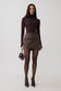 A model stands against a light gray studio background wearing a dark brown mock-neck long-sleeve top, a matching brown pleated wrap mini skirt, sheer black tights, and dark pointed-toe heels, while holding a small dark brown handbag.
