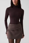 A model wearing a dark brown mock neck long-sleeve top and a brown pinstriped wrap mini skirt, photographed against a light gray studio background.