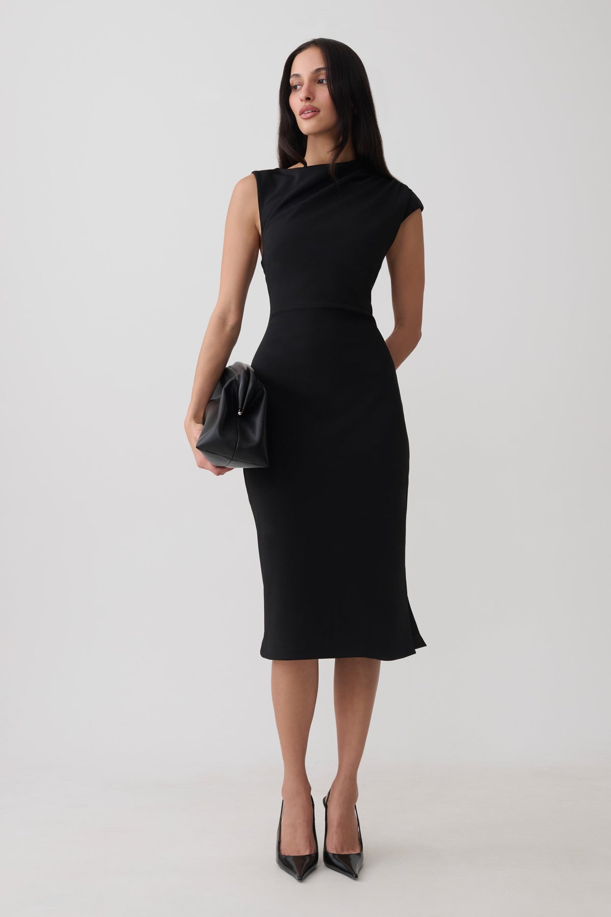 A woman modeling a black sleeveless midi dress with a high draped neckline, paired with black pointed-toe heels and a small black clutch, against a plain light gray studio background.