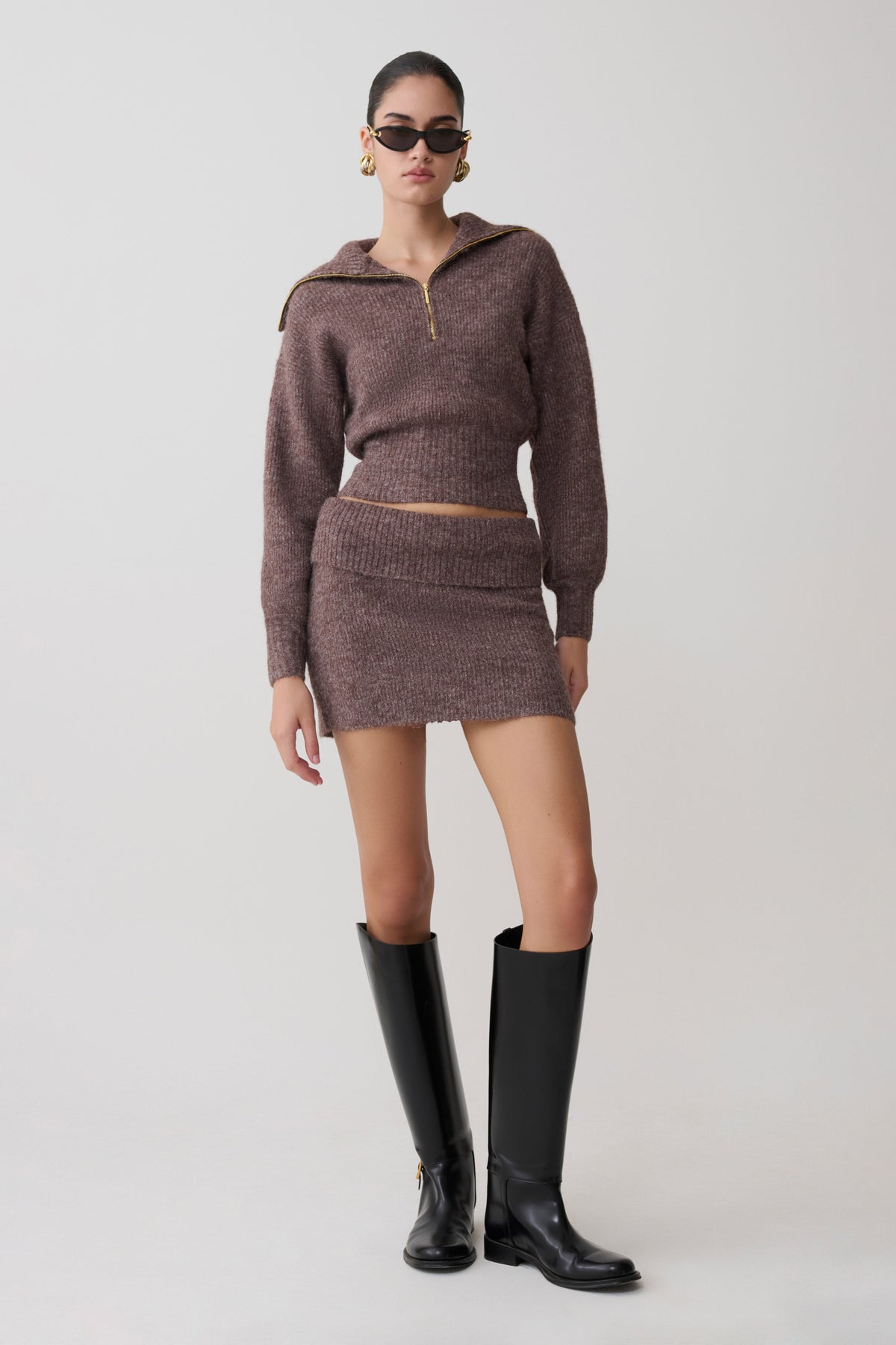 Woman wearing a brown knitted outfit with black knee-high boots on a white background.