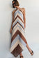 A woman modeling a halter-neck maxi dress with a chevron pattern in brown, beige, and light blue, paired with black sandals against a plain white wall.