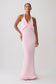 A woman modeling a light pink halter-neck maxi dress with a wrap-style bodice, photographed against a plain light gray studio background.