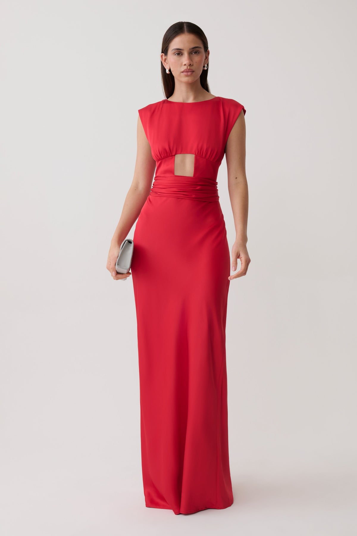 A woman modeling a bright red sleeveless maxi dress with a square neckline, a wrap-style cutout at the waist, and a fitted floor-length silhouette, photographed against a plain light gray studio background.