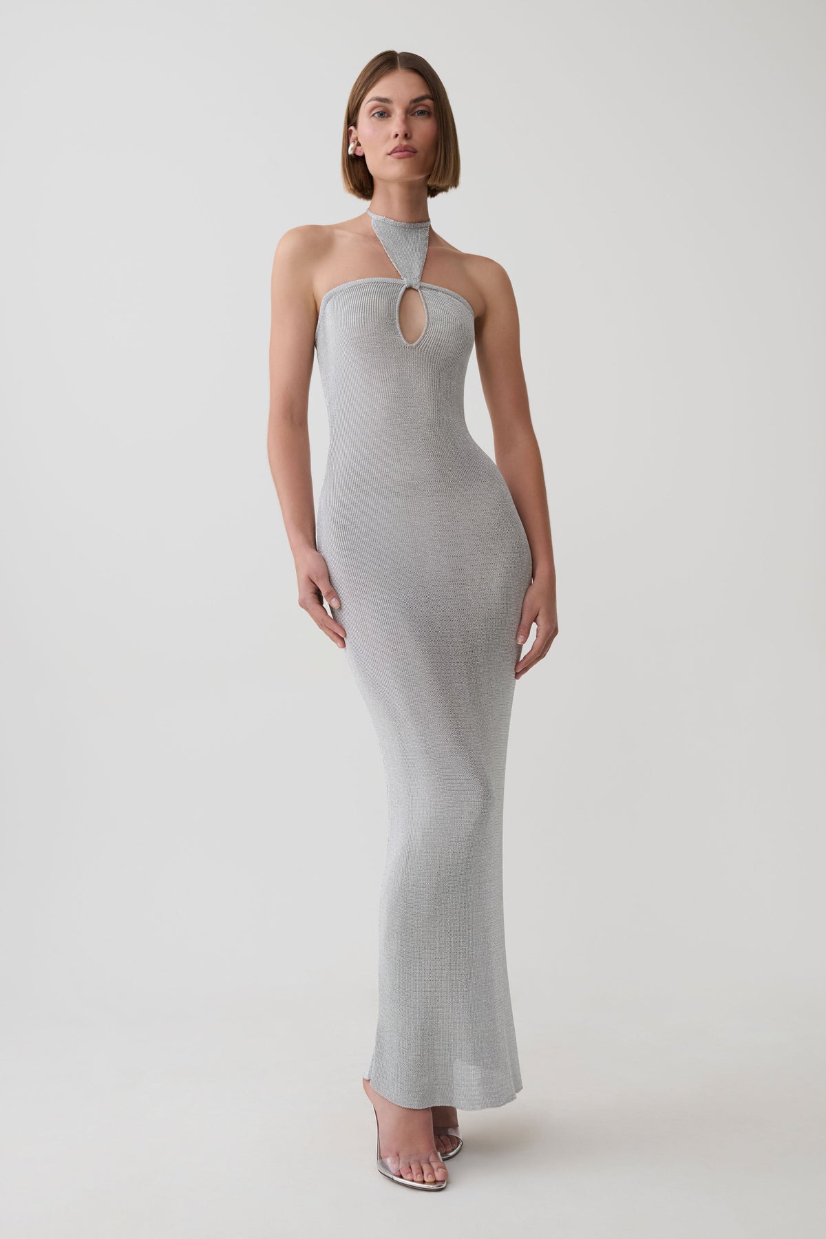 A woman modeling a light gray ribbed knit halter-neck maxi dress with a keyhole cutout at the chest, photographed against a seamless light gray studio background.