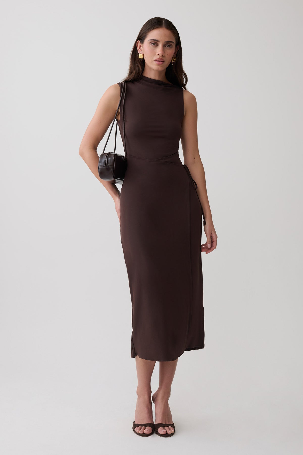 A woman modeling a sleeveless, high-neck, knee-length dark brown dress against a plain light gray studio background. She is carrying a small brown shoulder bag and wearing black heeled sandals.