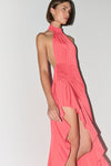 A woman modeling a coral pink halter-neck dress with a high-low ruffled hem and a side cutout, shown against a plain light gray studio background.