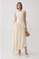 A woman models a sleeveless cream-colored midi dress with a structured bodice and a flowing skirt, holding a small woven straw handbag against a light gray studio background.