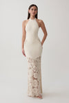 A woman modeling a sleeveless halter-neck maxi dress in cream with a textured crochet lace overlay on the lower half, set against a plain light gray studio background.