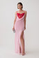 A model wearing a light pink slip-style maxi dress with a sweetheart neckline, sheer mesh bust, and a high thigh slit, paired with a bright red mesh corset mini dress underlayer and holding a small silver clutch against a plain light gray studio background.