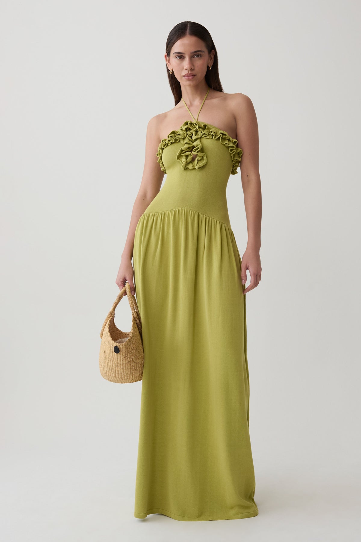 A woman modeling a strapless olive green maxi dress with a ruffled bodice and a matching fabric headband, holding a small woven straw handbag against a plain light gray studio background.