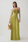 A woman modeling a strapless olive green maxi dress with a ruffled bodice and a matching fabric headband, holding a small woven straw handbag against a plain light gray studio background.