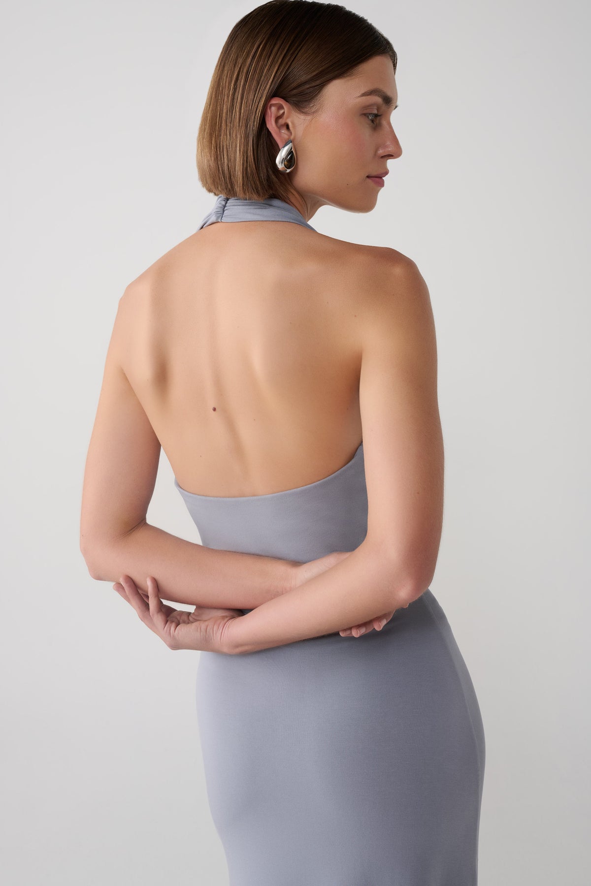 A woman wearing a sleeveless, high-neck, wide-leg jumpsuit in a dusty lavender-gray color, posing in a crouched position against a clean white wall.