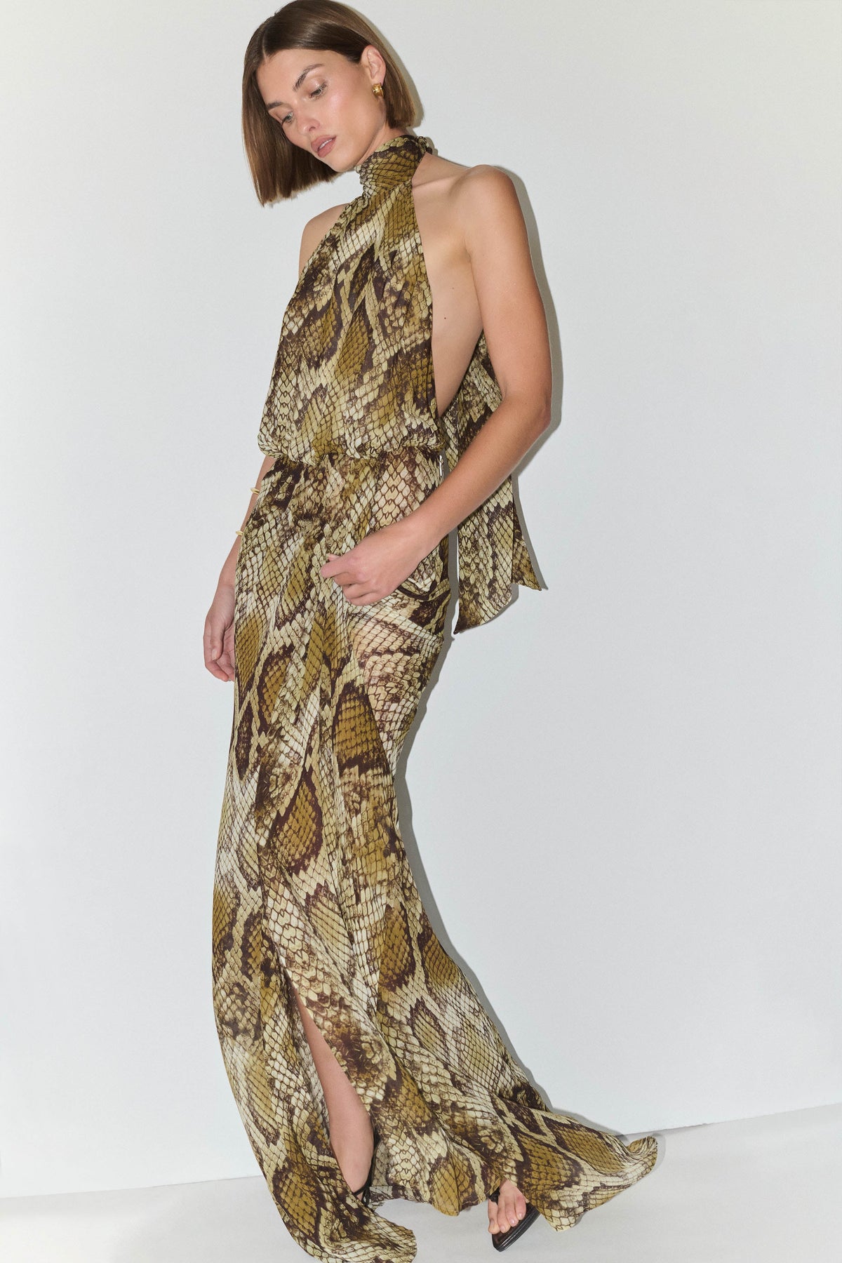 A woman modeling a sleeveless halter-neck maxi dress with a snakeskin print in beige, brown, and olive tones, featuring a high side slit and peplum waist detail, set against a plain white studio background.