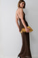 A woman wearing a brown spaghetti-strap maxi dress with a gold polka-dot pattern, holding a yellow woven clutch against a plain white studio background.