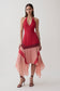 A woman modeling a sleeveless halter-neck midi dress with a deep V-neckline, featuring a solid red upper bodice and a layered, flowing peach-pink skirt with sheer side panels, set against a plain light gray studio background.