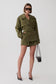 A model wearing an oversized olive green military-style trench jacket and matching shorts with a belt, styled with black pointed-toe heels and a small black shoulder bag against a light gray studio background.