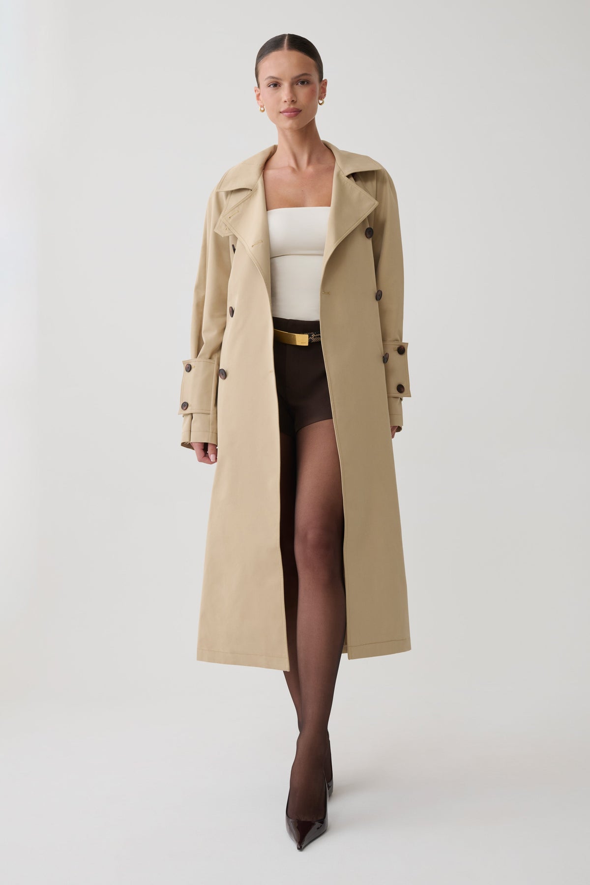 A model wearing a long beige trench coat over a white top, brown shorts, and dark brown tights, standing against a light gray studio background.