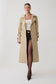 A model wearing a long beige trench coat over a white top, brown shorts, and dark brown tights, standing against a light gray studio background.