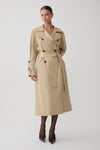 A woman modeling a mid-length beige trench coat with dark buttons, a matching waist belt, and dark tights with pointed heels, photographed against a plain light gray studio background.