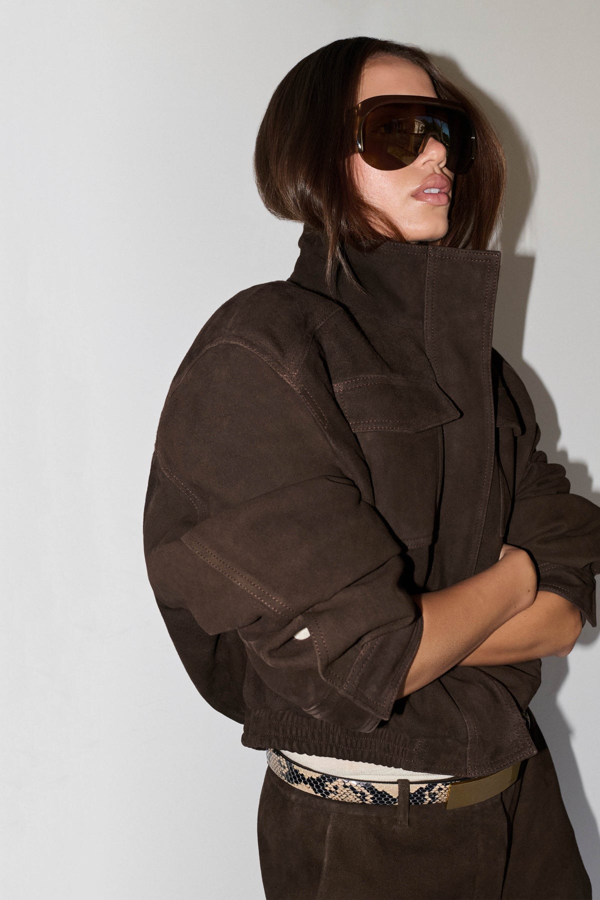 A model wearing a dark brown high-collar jacket made of textured suede-like fabric, styled with oversized dark sunglasses and a patterned belt, set against a plain light gray background.