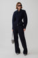 A model stands against a light gray studio background wearing a matching dark navy blue zip-up jacket and wide-leg trousers set, styled with black sunglasses, a small black-and-white zebra-patterned handbag, and black-and-white striped shoes.
