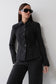 A woman wearing a black textured button-front jacket with structured shoulders and black pants, standing against a light gray wall.