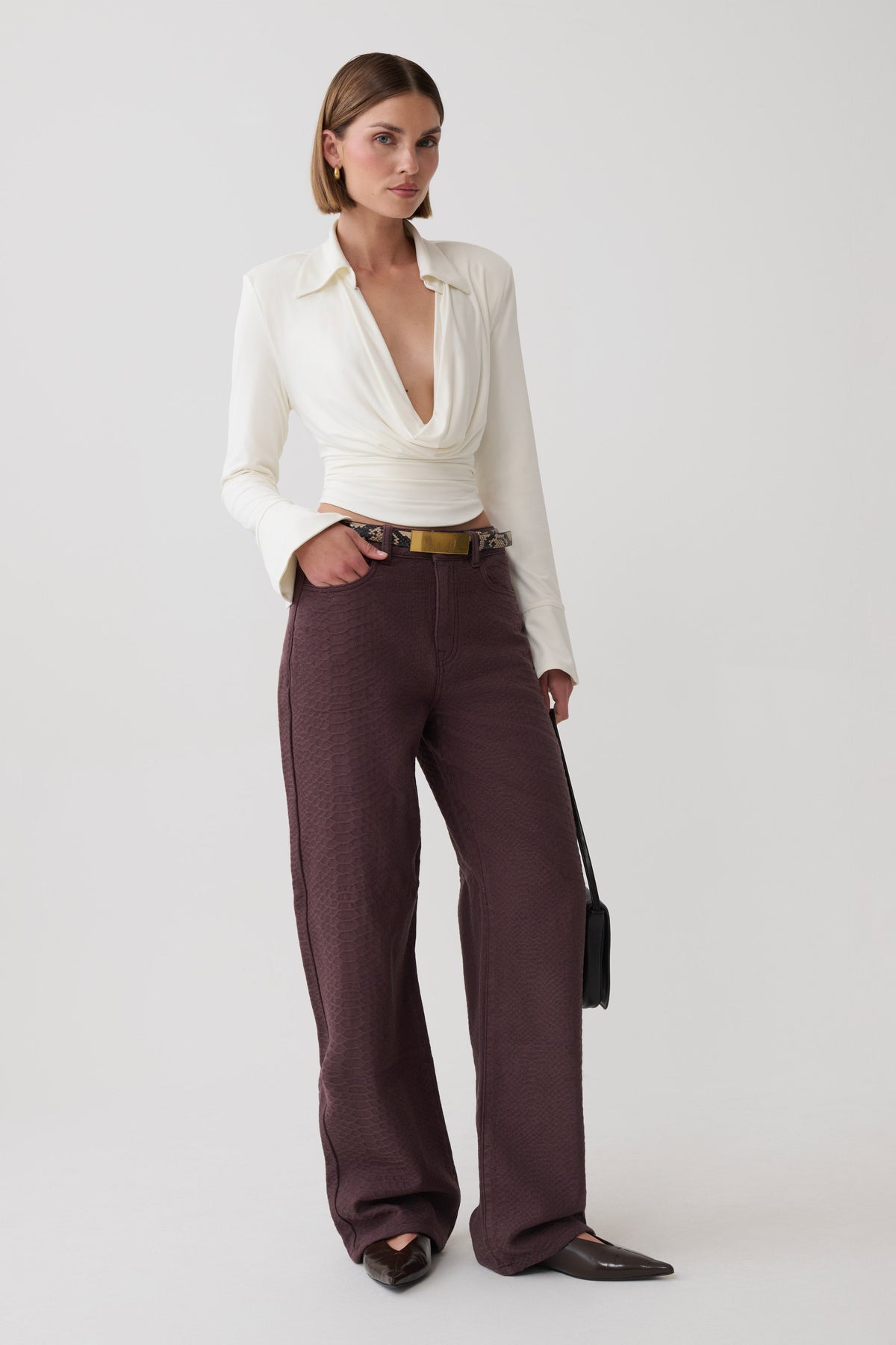 A model stands against a light gray studio background wearing a cream long-sleeve wrap-style blouse tucked into high-waisted, wide-leg dark brown trousers with a gold belt, paired with dark brown pointed flats and holding a small black handbag.