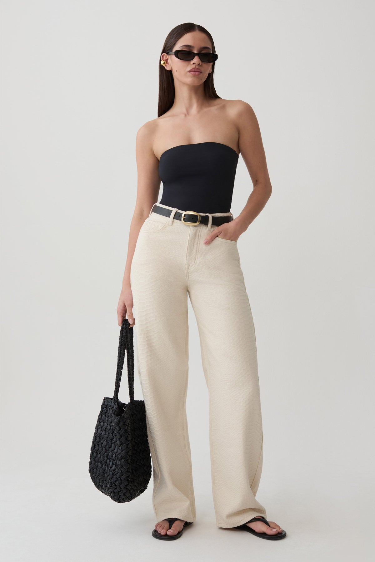 A woman modeling a black strapless bandeau top and cream wide-leg trousers, accessorized with a black woven tote bag, black sunglasses, and black sandals, against a light gray studio background.