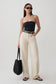 A woman modeling a black strapless bandeau top and cream wide-leg trousers, accessorized with a black woven tote bag, black sunglasses, and black sandals, against a light gray studio background.