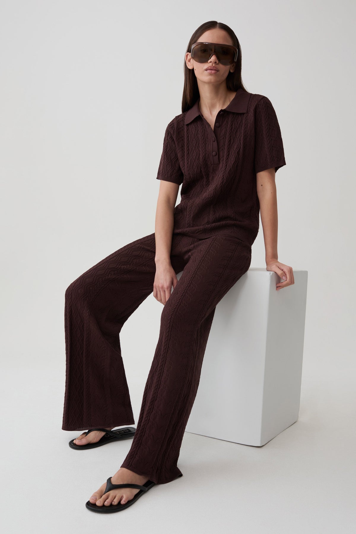 A woman wearing a matching dark brown short-sleeve collared top and wide-leg pants set, seated on a white block against a light gray studio background.