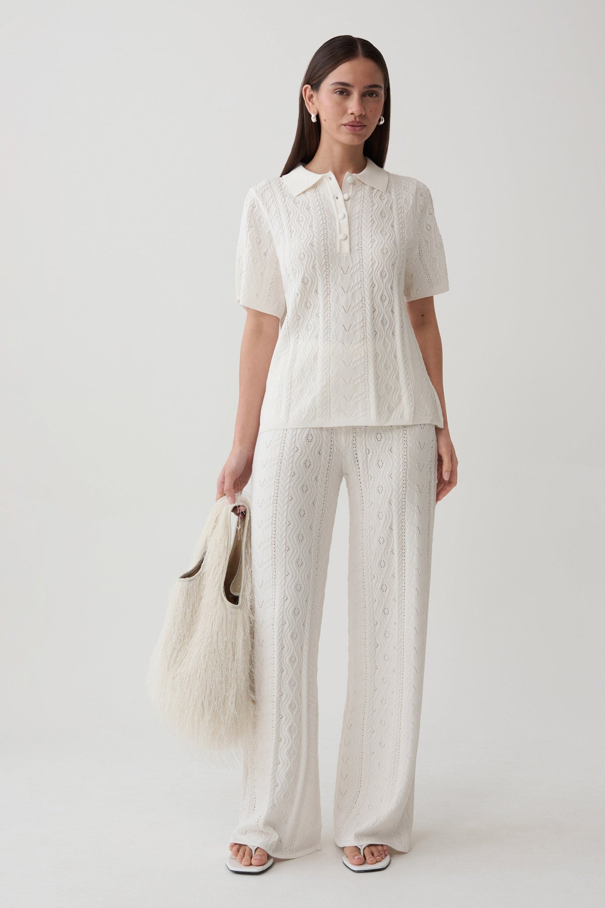 A woman modeling a matching two-piece outfit consisting of a short-sleeve collared blouse and wide-leg trousers in an off-white lace fabric, holding a fluffy cream tote bag against a plain light gray studio background.