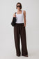 A model wearing a white tank top, dark brown wide-leg drawstring pants, black sunglasses, and carrying a black textured shoulder bag against a light gray studio background.
