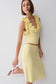 A woman models a pale yellow two-piece knit set consisting of a ruffled halter crop top and a matching midi skirt, paired with white heeled sandals against a plain light gray background.