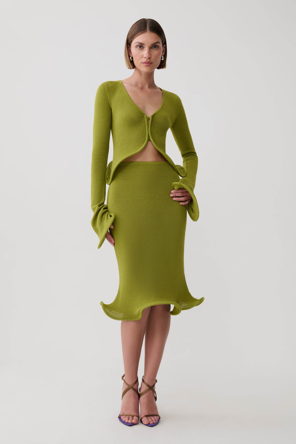 A model wearing a matching olive green knit two-piece set consisting of a long-sleeve cardigan with a front cutout and flared cuffs, and a fitted midi skirt with a ruffled hem, paired with brown strappy heels against a light gray studio background.