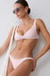 A woman modeling a light pink two-piece bikini set against a plain light gray background. She is wearing round sunglasses and a white textured shoulder bag.